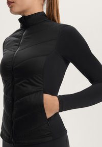 Black jacket featuring a fitted design with a zip closure, padded front, and long sleeves. Material is smooth with a textured, quilted pattern.