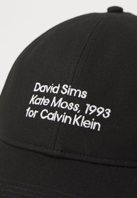 Fekete baseball sapka, fehér hímzett felirattal: "David Sims Kate Moss, 1993 a Calvin Klein számára" a frontpanelen.