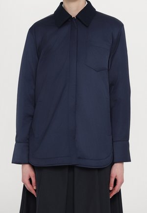 Giubbotto trapuntato blu navy con colletto a punta, chiusura frontale con bottoni e taschino sul petto. Il design presenta maniche lunghe con polsini più larghi.
