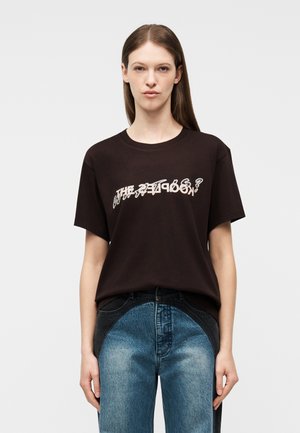 Kvinde med glat brunt hår, iført en sort t-shirt med grafik og blå-sorte tofarvede jeans, stående foran en ensfarvet hvid baggrund.