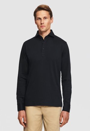 Mężczyzna w czarnej koszulce polo z długim rękawem i beżowych spodniach, stojący na jednolitym jasnoszarym tle.