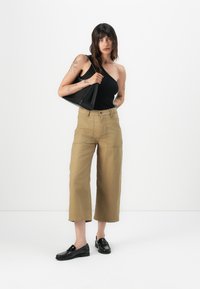 Pantaloni larghi color marrone chiaro con lunghezza cropped, dotati di tasche frontali e chiusura con bottone, abbinati a un top nero a spalla unica e mocassini.