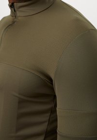 Haut de sport vert olive à manches longues avec un col montant, présentant une texture côtelée, une fermeture éclair et une conception d'épaule sans coutures.