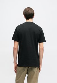 Melnas kokvilnas T krekls ar klasisku apaļu kakla izgriezumu un īsām piedurknēm; attēlots no aizmugures, ar brīvu griezumu un gludu tekstūru.