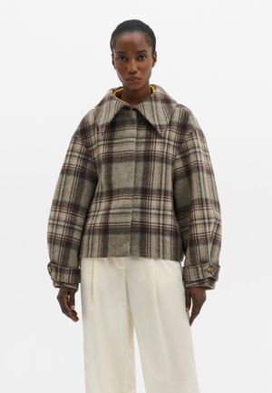 Frau trägt eine grau-braune karierte Wolljacke mit weitem Kragen und cremefarbenen, weiten Hosen, steht vor einem schlichten weißen Hintergrund.