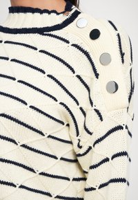 Kreminė megzta džemperis su tamsiai mėlynu geometriniu raštu. Turintis iškilų apykaklę ir pečių sagas. Minkšta tekstūra ir struktūruota dizaino.