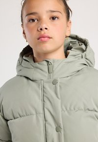 Joven con ojos marrones y pendientes, vistiendo una chaqueta acolchada de color verde claro con capucha y cremallera, mirando al frente con calma.