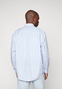 Camisa de manga larga azul claro con rayas verticales blancas, que presenta un cuello de botón y un dobladillo curvado, hecha de una tela suave.