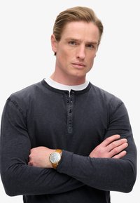 Dunkelblaues Langarm-Henley-Shirt mit einem weißen Kragen, ausgestattet mit Knöpfen am Hals. Am Handgelenk wird eine braune Lederuhr getragen.
