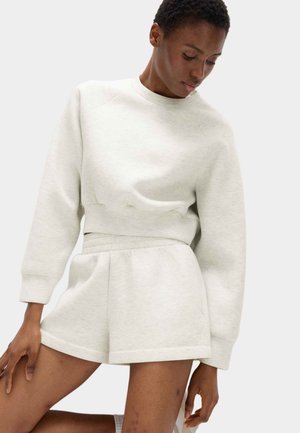 Person iført lyse beige, cropped sweatshirt med lange ærmer og matchende shorts med høj talje, kigger nedad mod en ensfarvet baggrund.