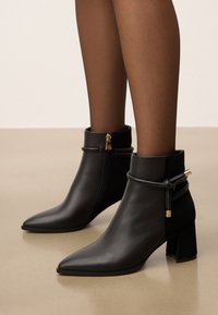 Fekete hegyes orrú bokacsizmában, blokktalpon, oldalsó cipzárral és díszes, körbetekert pántokkal, átlátszó fekete harisnyával párosítva.