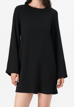 Frau trägt ein locker sitzendes schwarzes Mini-Kleid mit langen Ärmeln und rundem Ausschnitt, steht vor einem schlichten weißen Hintergrund.