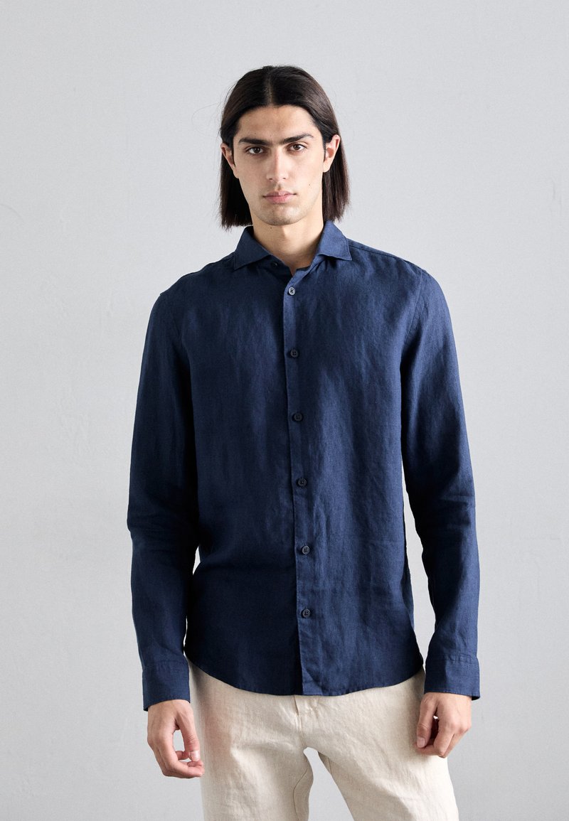 Camisa de lino de manga larga azul marino con cuello abotonado y dobladillo recto. Presenta una textura en la tela y un bolsillo en la parte delantera.
