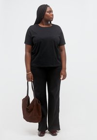 Γυναίκα με πλεξούδες που φοράει μαύρο μπλουζάκι και παντελόνι, κρατώντας καφέ τσάντα ώμου, κοιτάζοντας προς τα δεξιά της, μπροστά σε απλό ανοιχτό φόντο.