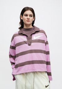 Νεαρός που φοράει μπλούζα Nike με λωρίδες μωβ και καφέ και ανοιχτό παντελόνι με το λογότυπο της Nike, κοιτάζοντας ελαφρώς προς το πλάι πάνω σε λευκό φόντο.