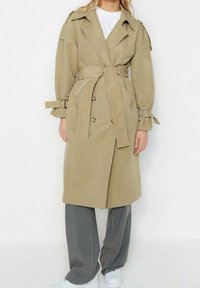 Kobieta ubrana w beżowy płaszcz trencz z paskiem, nosząca szare spodnie o szerokich nogawkach i białe sneakersy, stojąca na tle gładkiej białej ściany.