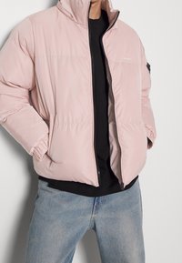 Rosa puffjacka med hög krage, dragkedjestängning, sidofickor och elastiska muddar. Bärs över en svart skjorta och matchas med blå jeans.