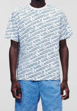 Mężczyzna ubrany w białą, krótko rękawową koszulę i niebieskie spodnie, obie części garderoby ozdobione powtarzającym się wzorem z napisem "Karl Lagerfeld".