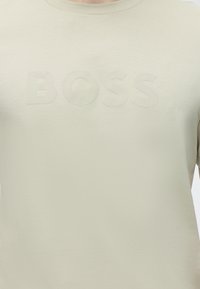 T-shirt beige à col rond avec le lettrage embossé « BOSS » sur la poitrine, porté par une personne visible des épaules jusqu'au milieu du torse.