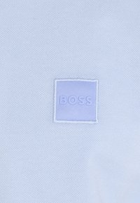 Svetlomodrá látka má prišitou štvorcovou patch s embosovaným textom "BOSS" v strede. Textúra je hladká s jemným tkaním.