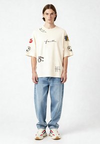 Krema krāsa oversized T-krekls ar dažādiem krāsainiem grafiskajiem attēliem, tostarp "ticība", lūpām un sejām, pāris ar gaiši ziliem platām biksēm un sporta apaviem.