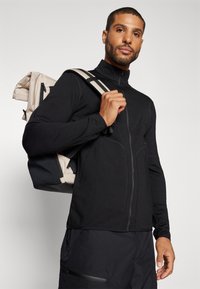 Veste noire zippée avec col haut, design ajusté, associée à un sac à dos à carreaux de couleur claire avec des accents noirs et des bretelles réglables.