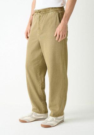 Mann trägt beige locker sitzende Hose mit elastischem Bund und weiße Sneakers mit braunen Sohlen, steht vor schlichtem weißem Hintergrund.