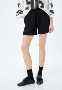 Pantaloni corti neri in cotone con vita elastica e cordino, caratterizzati da una texture liscia e una vestibilità casual, abbinati a sneakers nere e calze.