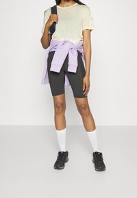 Abbigliamento sportivo che include una t-shirt beige chiaro, shorts da ciclismo neri, una felpa lavanda legata in vita e sneakers nere con calzini bianchi.
