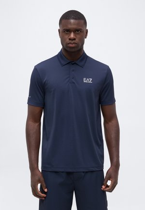 Homme portant un polo bleu marine EA7 Emporio Armani et un pantalon assorti, debout devant un fond blanc uni.