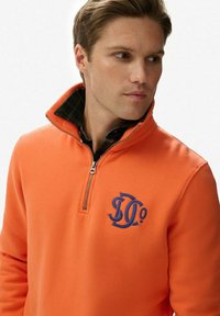 Pulóver de forro polar naranja con cuello con cremallera, que presenta un logo azul en el pecho y un cuello a cuadros verde debajo. Textura suave.