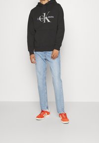 Sudadera negra con capucha con el logotipo "Calvin Klein Jeans", combinada con jeans de color azul claro y zapatillas de ante naranjas con acentos blancos.