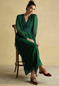 Długa zielona sukienka z geometrycznym wzorem, przezroczyste rękawy i głęboki dekolt w serek. Połączona z bordowymi butami na paski, na drewnianym krześle.