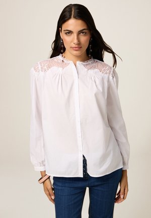 Hvid skjorte med knapper og blonde detaljer ved skuldrene, lange ærmer og en let løs pasform, kombineret med mørkeblå jeans.