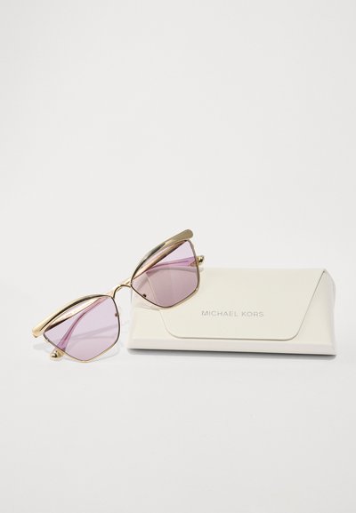 Lunettes de soleil Michael Kors avec verres roses, monture en métal doré et forme originale en œil de chat, reposant sur un étui blanc texturé arborant le logo.