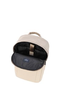 Mochila beige con un diseño de parte superior redondeada, exterior de tela, interior forrado en negro y un compartimento con cremallera. Cuenta con un asa y correas ajustables.