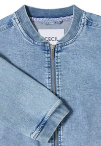 Veste bomber en denim bleu clair avec col et poignets côtelés, fermeture éclair à l'avant et étiquette de marque visible à l'intérieur du col.