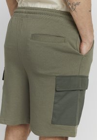 Shorts en coton vert olive avec poches cargo latérales, une poche arrière et une taille élastique ; texture lisse, coupe décontractée.