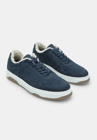 Jūras spēku zamšas tenisa kurpes ar baltām gumijas zoles, ar vilnas oderi un teksturētu virsējo daļu. Iekļauj auklu aizdari un sānu šuvju apdari.