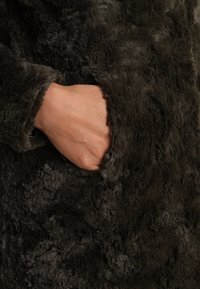 Luddigt mörkgrått tyg med en mjuk textur; en hand vilar i en sidoficka och visar upp rockens mjuka material och designdetaljer.