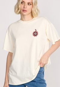T-shirt oversize couleur crème en coton doux, avec un graphique floral rose et noir sur la poitrine gauche. Coupe décontractée avec des manches courtes.