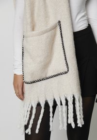 Châle en tricot couleur crème avec un motif en chevrons texturé, présentant une grande poche et des bords à franges soulignés d'un liseré noir.