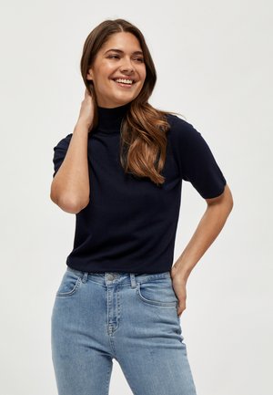 Nasmejana ženska z dolgimi rjavimi lasmi nosi mornarsko modro kratkoročno majico z visokim ovratnikom in svetlo modre kavbojke z visokim pasom, pózira z roko za vratom.