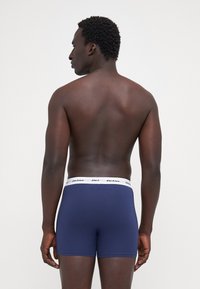 Marineblaue Boxershorts mit einem weißen elastischen Bund, auf dem der Markenname steht. Glatte Textur, körpernahe Passform, Beinform bis zur Mitte des Oberschenkels.