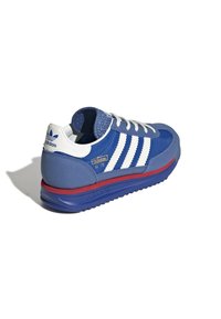 Baskets de sport bleues avec des rayures blanches et des accents gris. Présentent un talon texturé, une languette rembourrée et une semelle intermédiaire rouge. Style rétro classique.