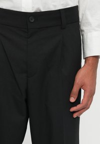 Pantalones negros a medida que presentan un cierre de botón único y detalles de pliegues, hechos de una tela texturizada con sutiles rayas. 