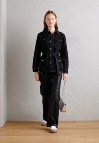 Giacca di jeans nera con tasche anteriori e una cintura, abbinata a pantaloni neri. Il modello tiene in mano una borsa con accenti argentati e indossa sneakers bianche.