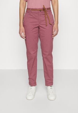 Femme portant un pantalon slim mauve avec une ceinture tressée marron et des baskets blanches Adidas, debout devant un fond uni.