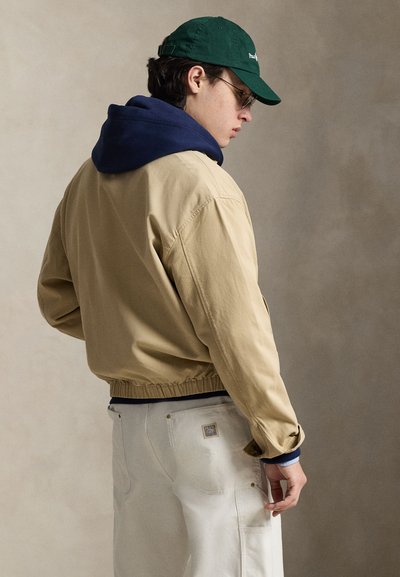 Μπεζ μπουφάν bomber με σκούρο μπλε κουκούλα, ελαστική μέση και λεπτομέρειες στις μανσέτες. Φορεμένο με εκρού παντελόνι που διαθέτει τσέπη και χρυσές μεταλλικές λεπτομέρειες.