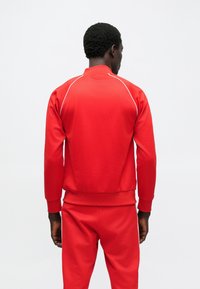 Fato de treino vermelho com acabamento texturizado, apresentando gola com fecho de correr, mangas raglan e detalhes em piping branco ao longo dos ombros e costuras.